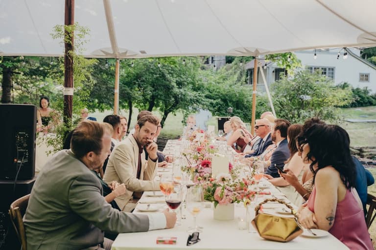 Guests seated at a long tented reception table strewn with flowers