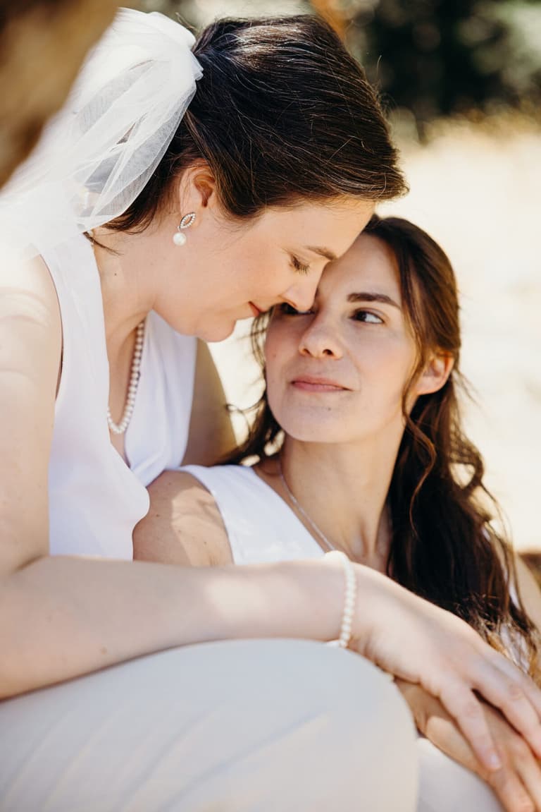 Two brides in white leaning close in warm sunlight