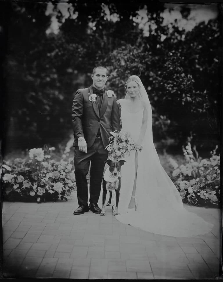 Newlyweds with their dog on a flower-lined brick path