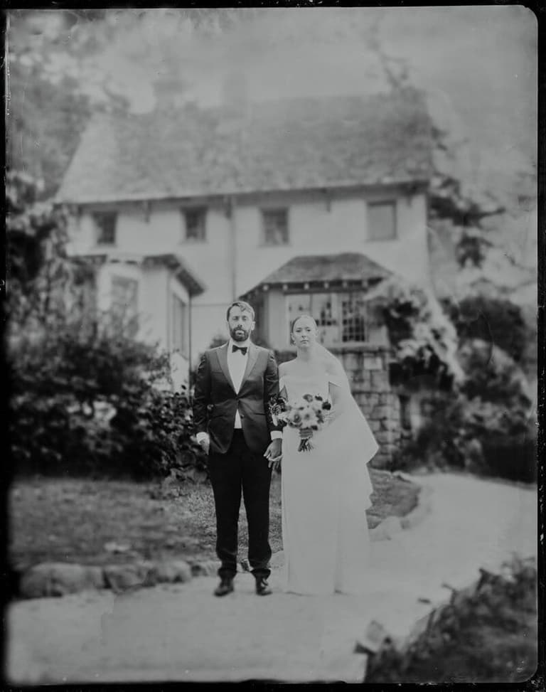 Bride and groom walking arm-in-arm past a white cottage, holding flowers in black-and-white film photography at their Hastings House wedding on Salt Spring Island