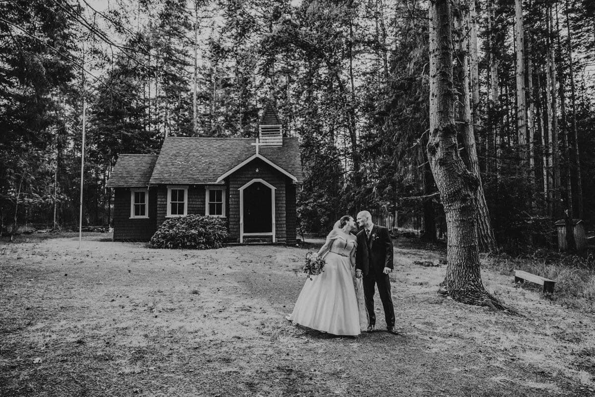 Newlyweds beside a tiny forest chapel in black and white