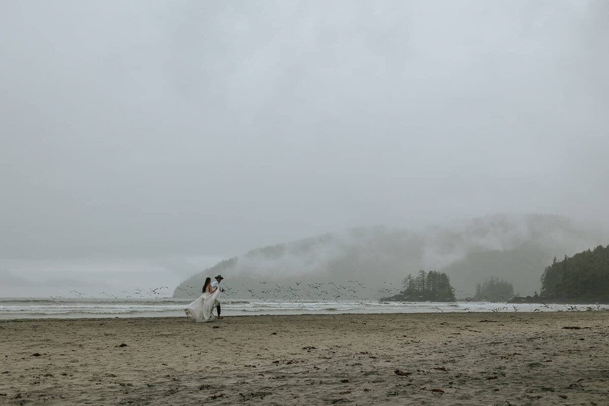Bride and groom crossing a misty central Vancouver Island beach as seabirds fly