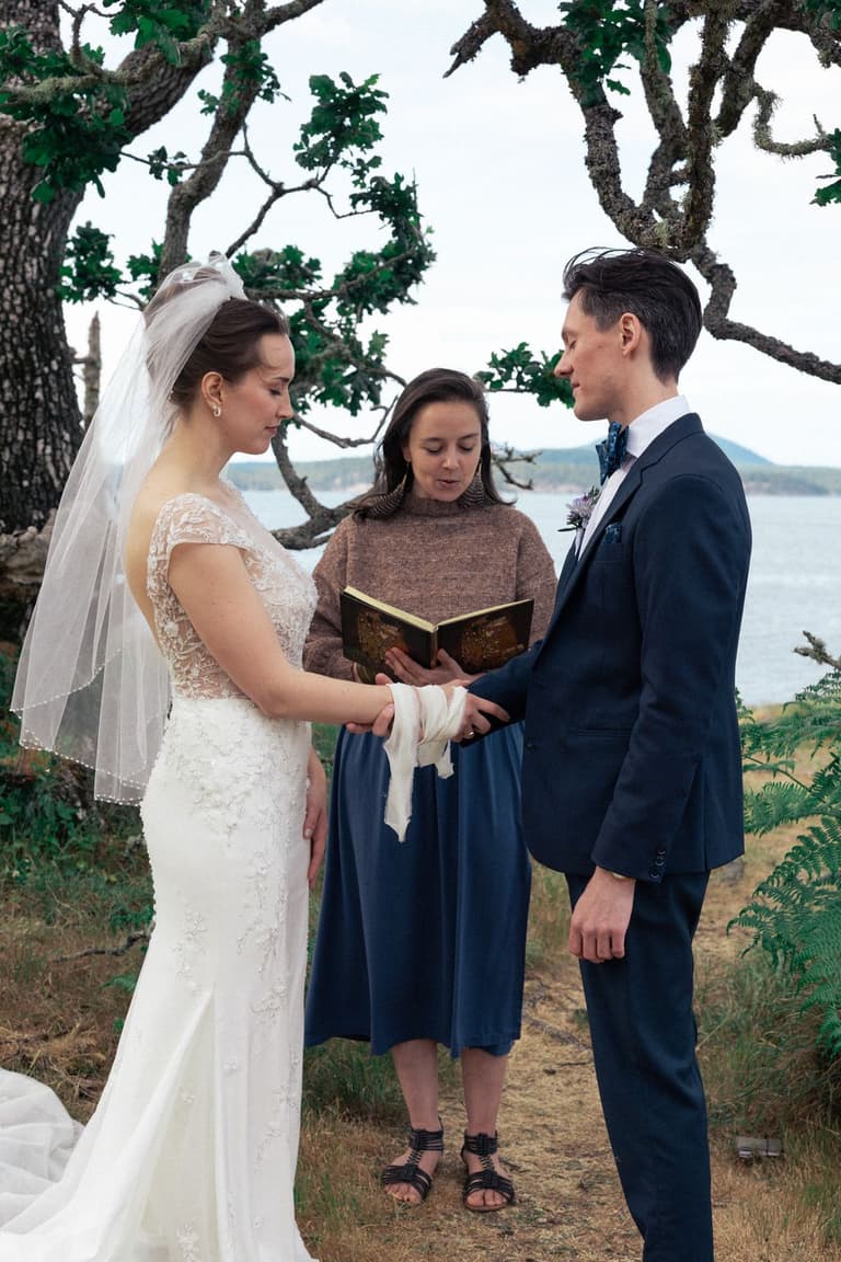 Officiant reads vows from leather-bound book as bride and groom hold hands beneath gnarled tree overlooking water