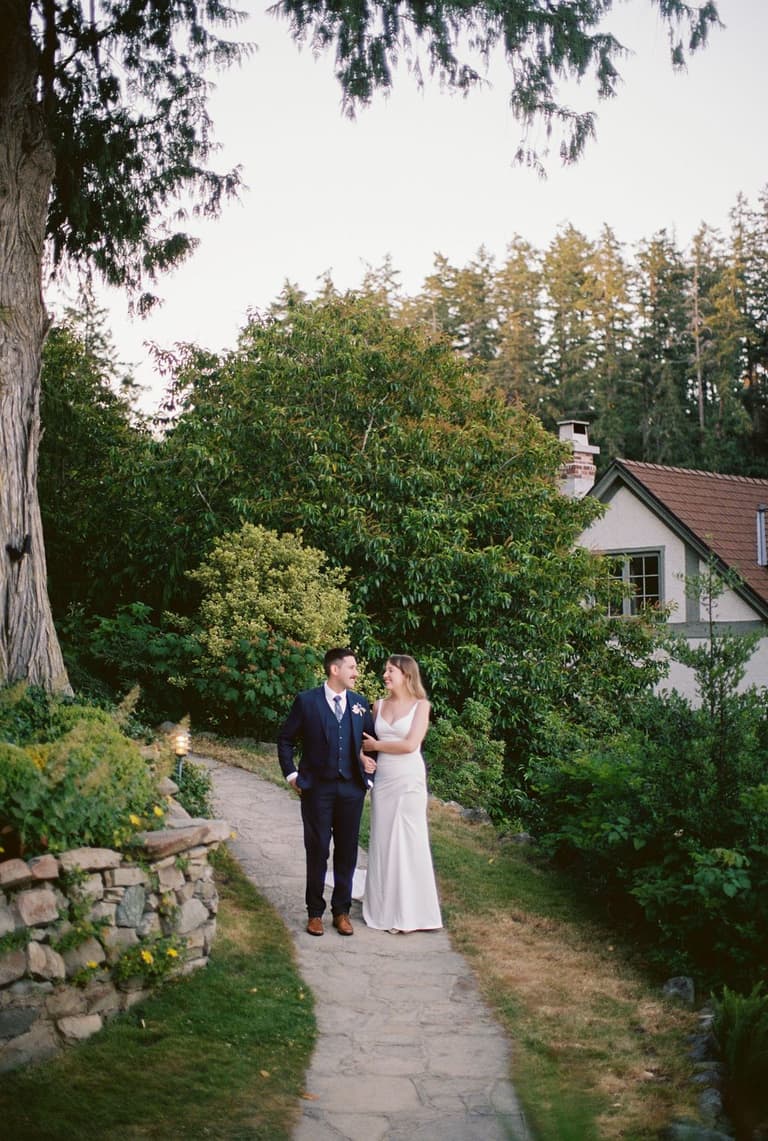 Bride and groom walking arm-in-arm on a garden path lined with lush green foliage and hanging wisteria