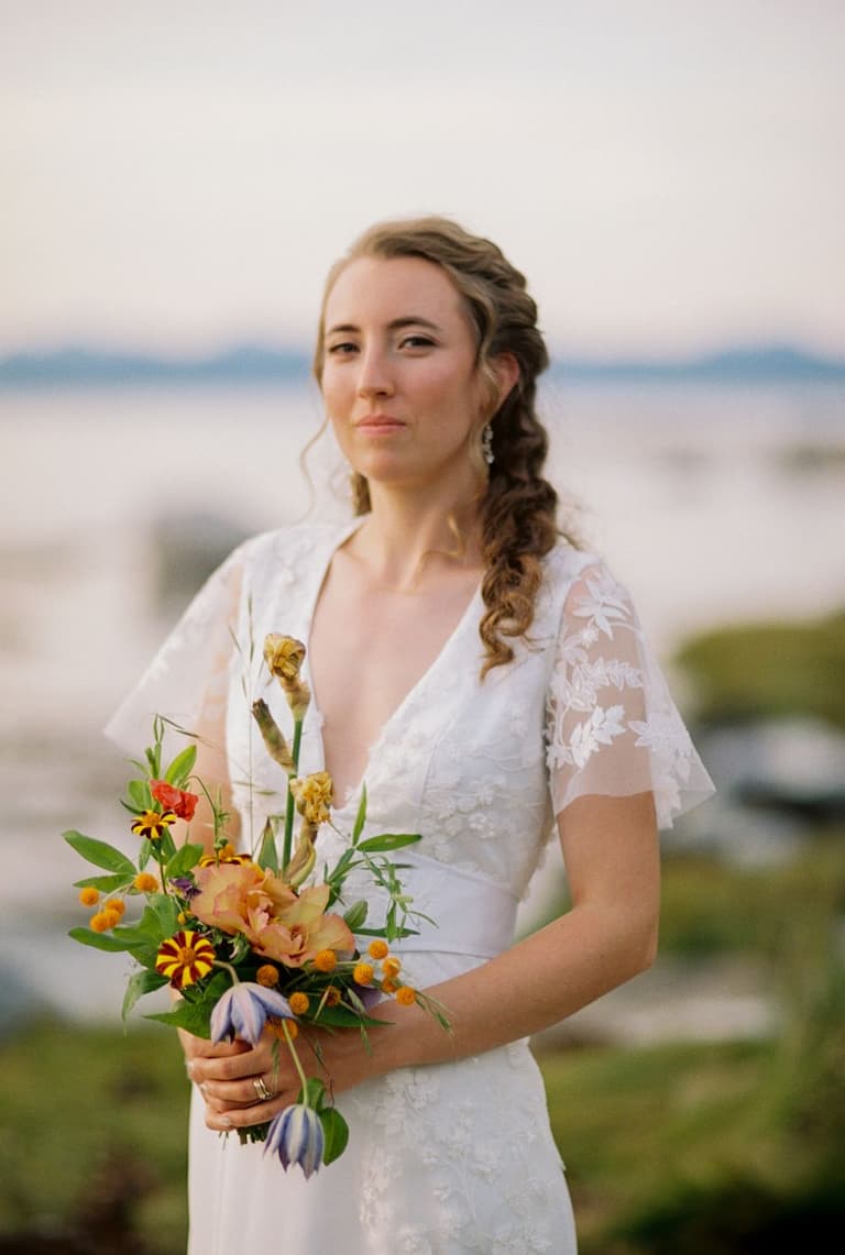 Bride in white lace dress holding wildflower bouquet gazes toward ocean under soft evening light on Vancouver Island.